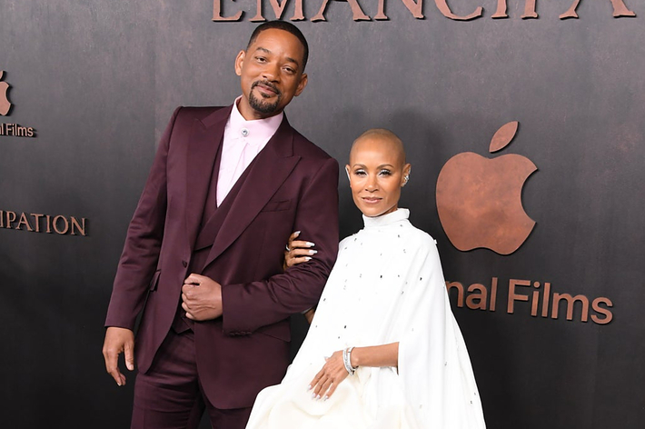 Will Smith and Jada Pinkett Smith posing together; he's in a burgundy suit, she's in a white dress with dots