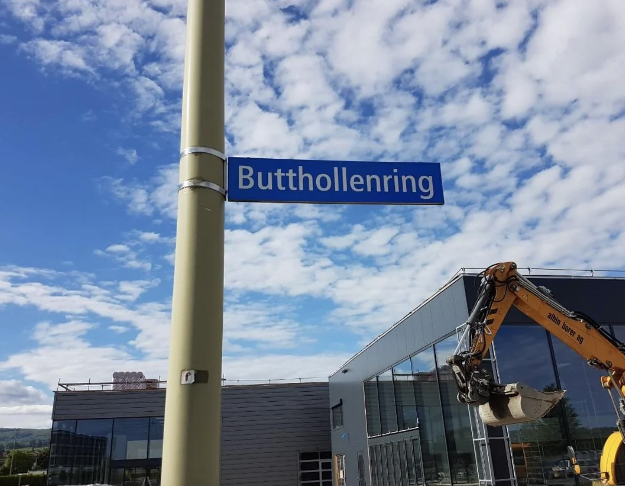 Sign reading &#x27;Butthollenring&#x27; above a street, with a clear sky, construction equipment, and a building in the background