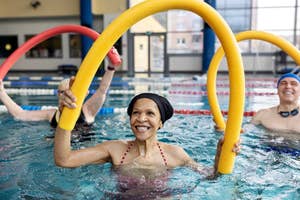Senior woman with a swim cap smiling in a pool, exercising with a yellow foam noodle, man in background also with noodle