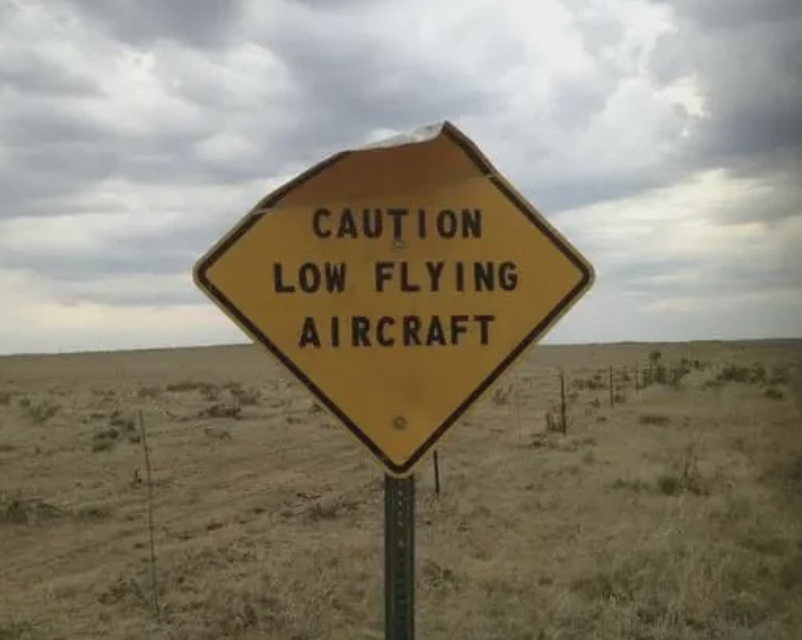 Warning sign reads &quot;CAUTION LOW FLYING AIRCRAFT&quot; with a flat landscape in the background