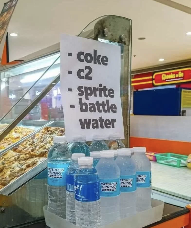 Sign displaying drink options: coke, C2, sprite, battle water, beside bottled waters at a food stall
