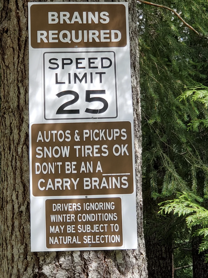 Sign on a tree reads &quot;BRAINS REQUIRED&quot;, &quot;SPEED LIMIT 25&quot;, &quot;AUTOS PICKUPS SNOW TIRES OK DON&#x27;T BE A CARRY A BRAINS&quot;, and warning about ignoring winter conditions