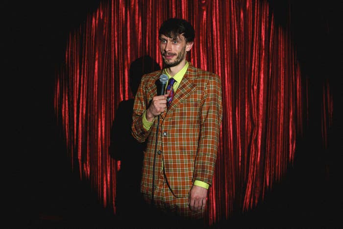 Richard Gadd in a plaid suit stands onstage holding a microphone in front of curtain