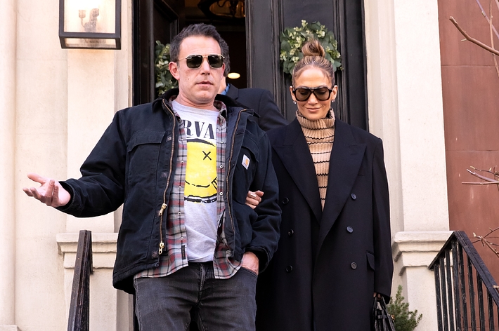 Two individuals exit a building; the person on the left wears a graphic tee and jacket, the right in a turtleneck and coat