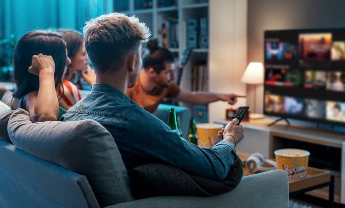 Friends sitting on a couch watching TV, a man holds a remote, and the screen displays a movie selection interface