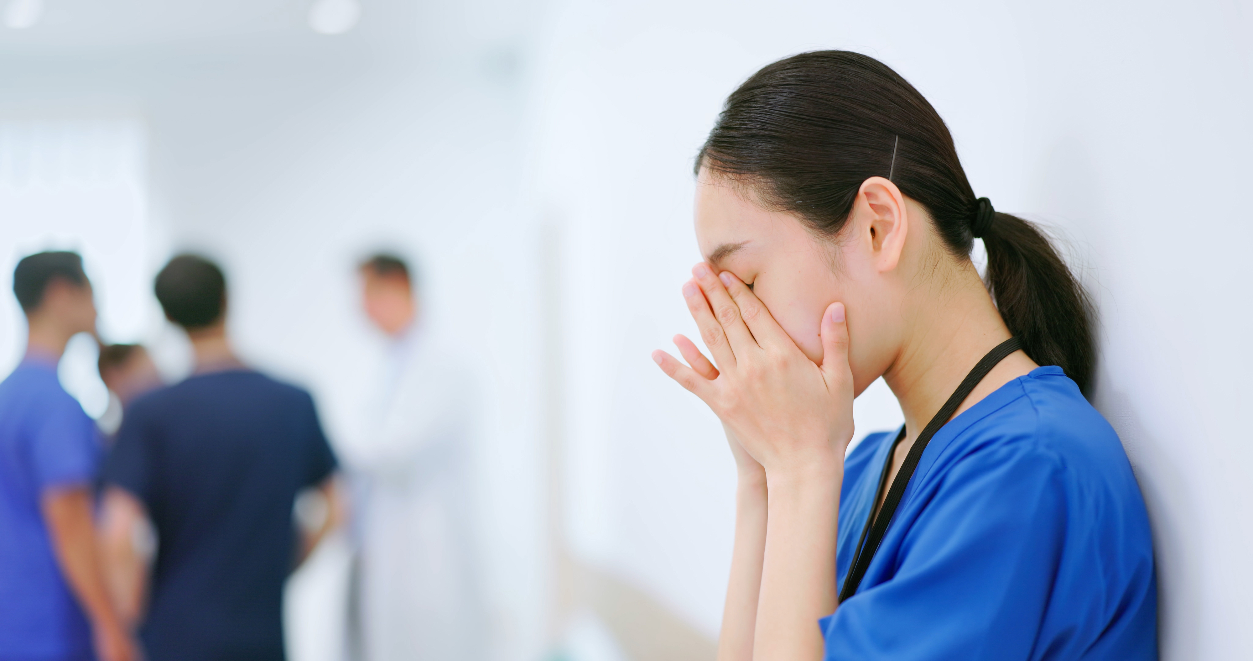 Woman in scrubs appears stressed with hand on forehead, colleagues in background