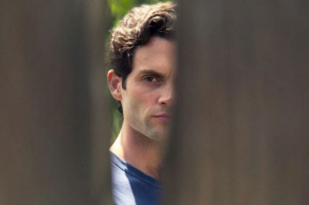 A man is partially visible, peering through a gap in a wooden fence