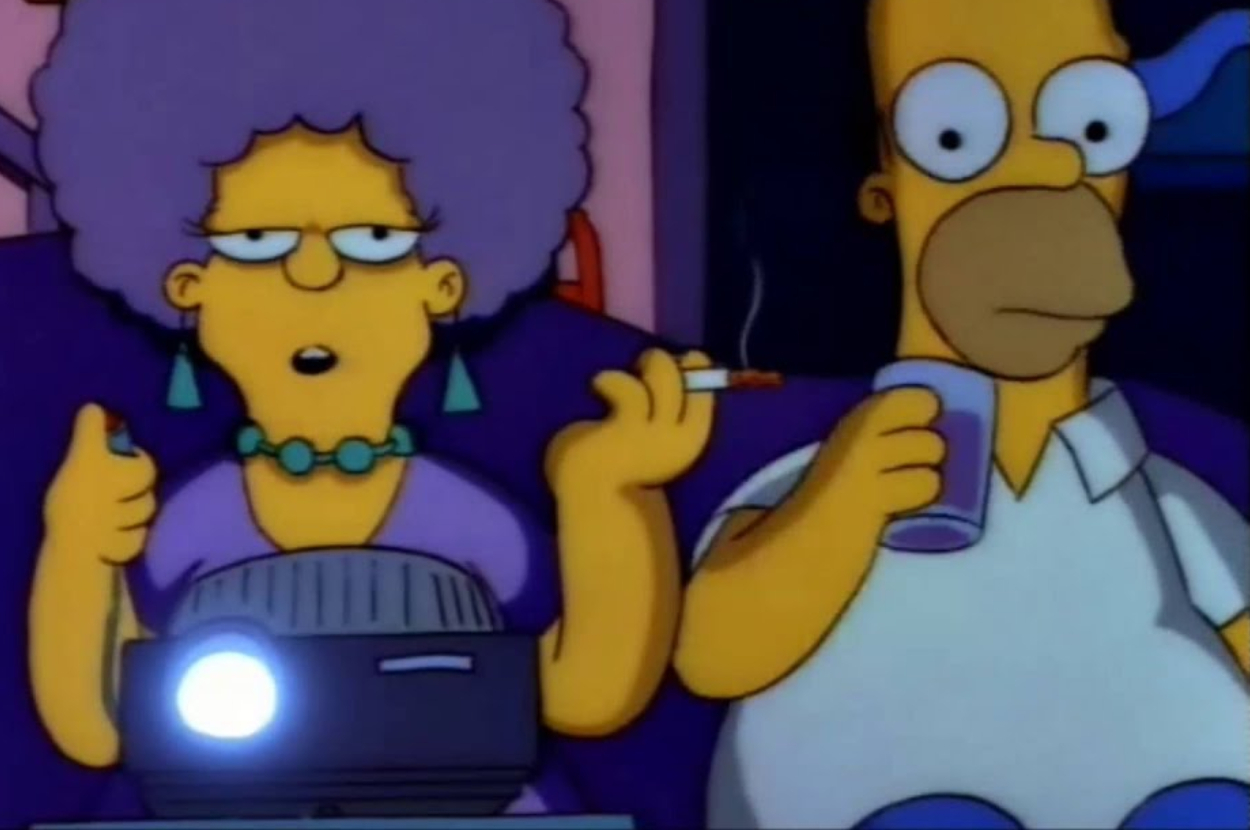 Patty Bouvier and Homer Simpson are sitting on a sofa. Patty is holding a lit cigarette and a projector remote, while Homer is holding a beverage cup