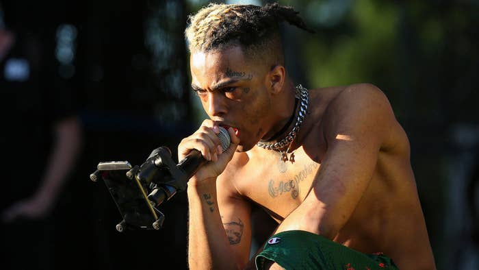 A shirtless performer with tattoos, a choker, and distinct hair sings into a microphone on stage while holding a phone