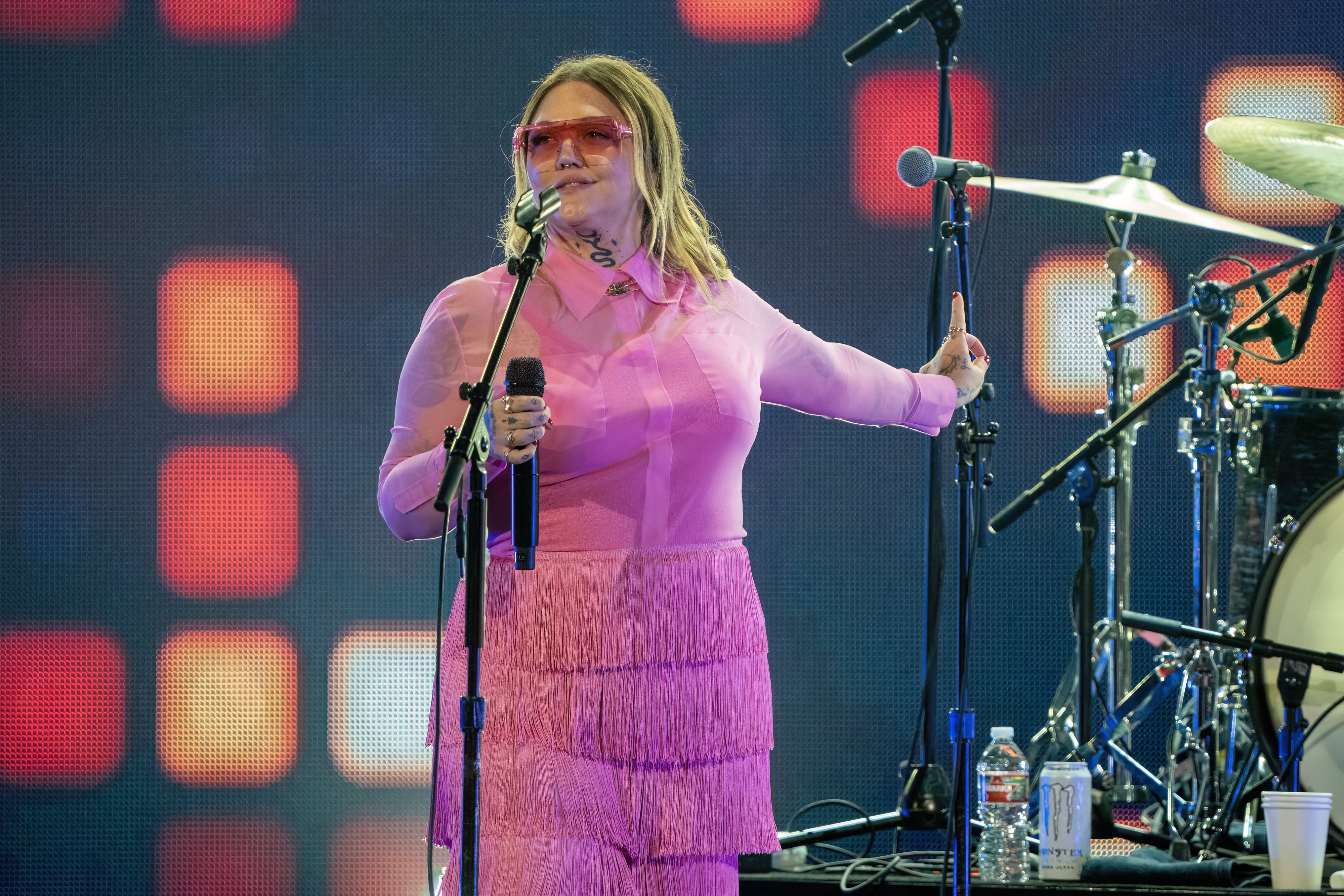 Elle King performs onstage, wearing a fringed outfit and sunglasses, holding a microphone