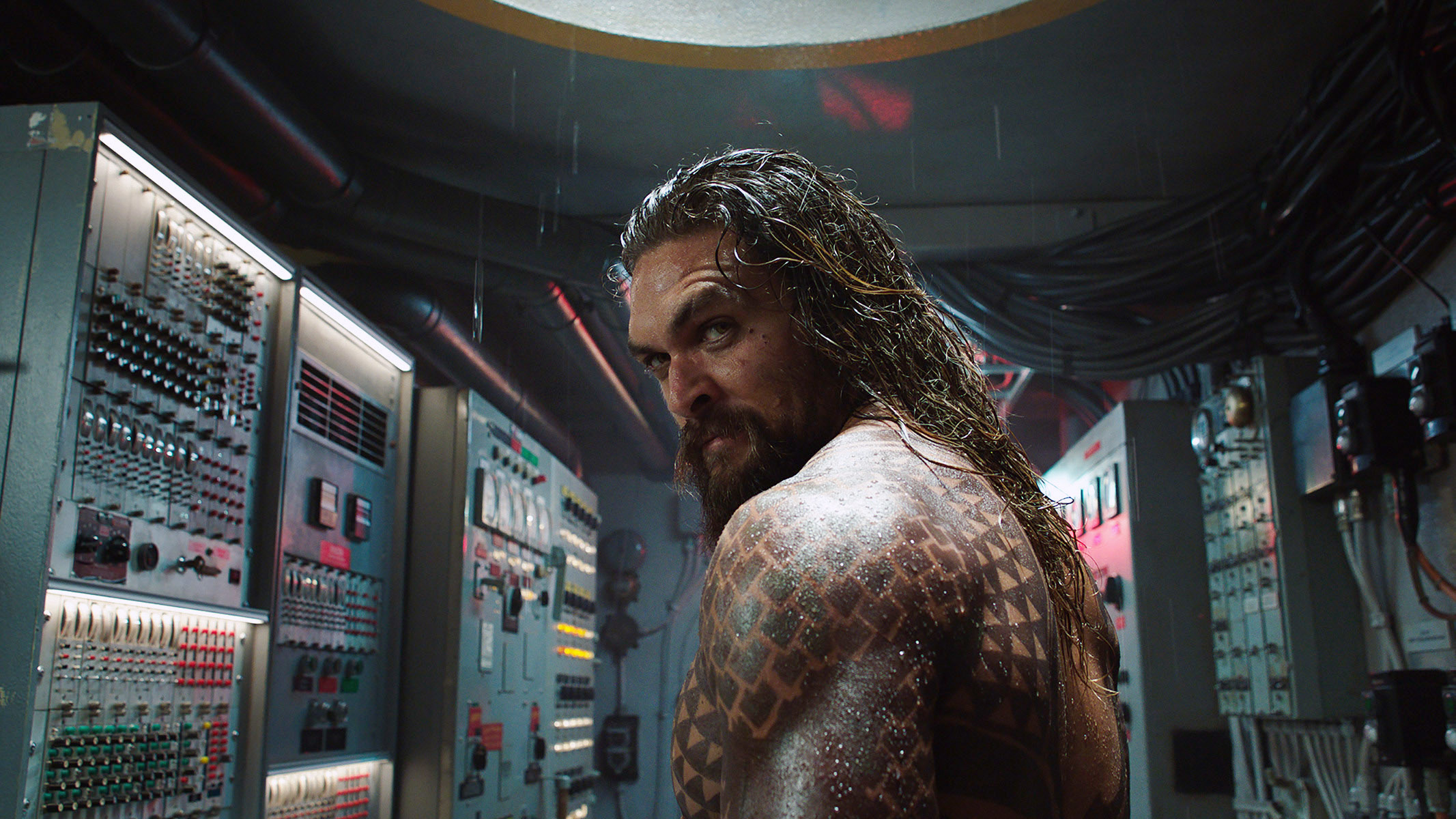 Jason Momoa, shirtless, with long wet hair and tattoos, stands in a control room filled with machinery and electrical panels, in a scene