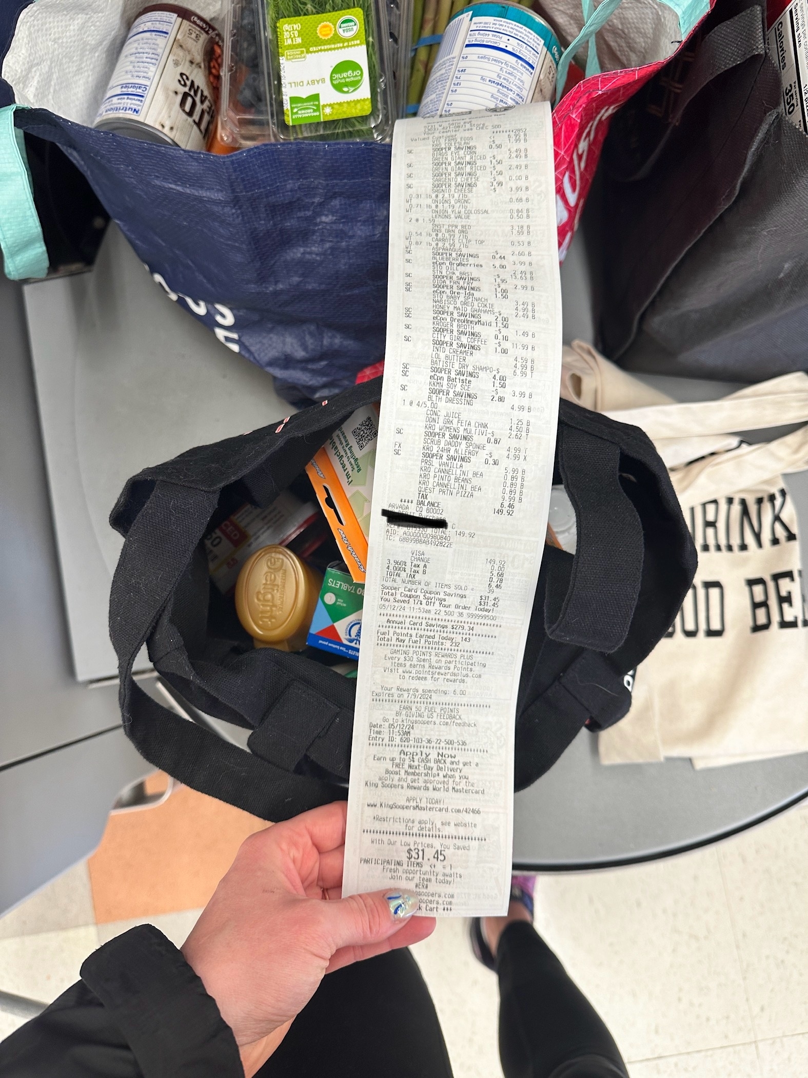 A hand holding a long shopping receipt over bags of groceries, including a can, packaged item, and other food products