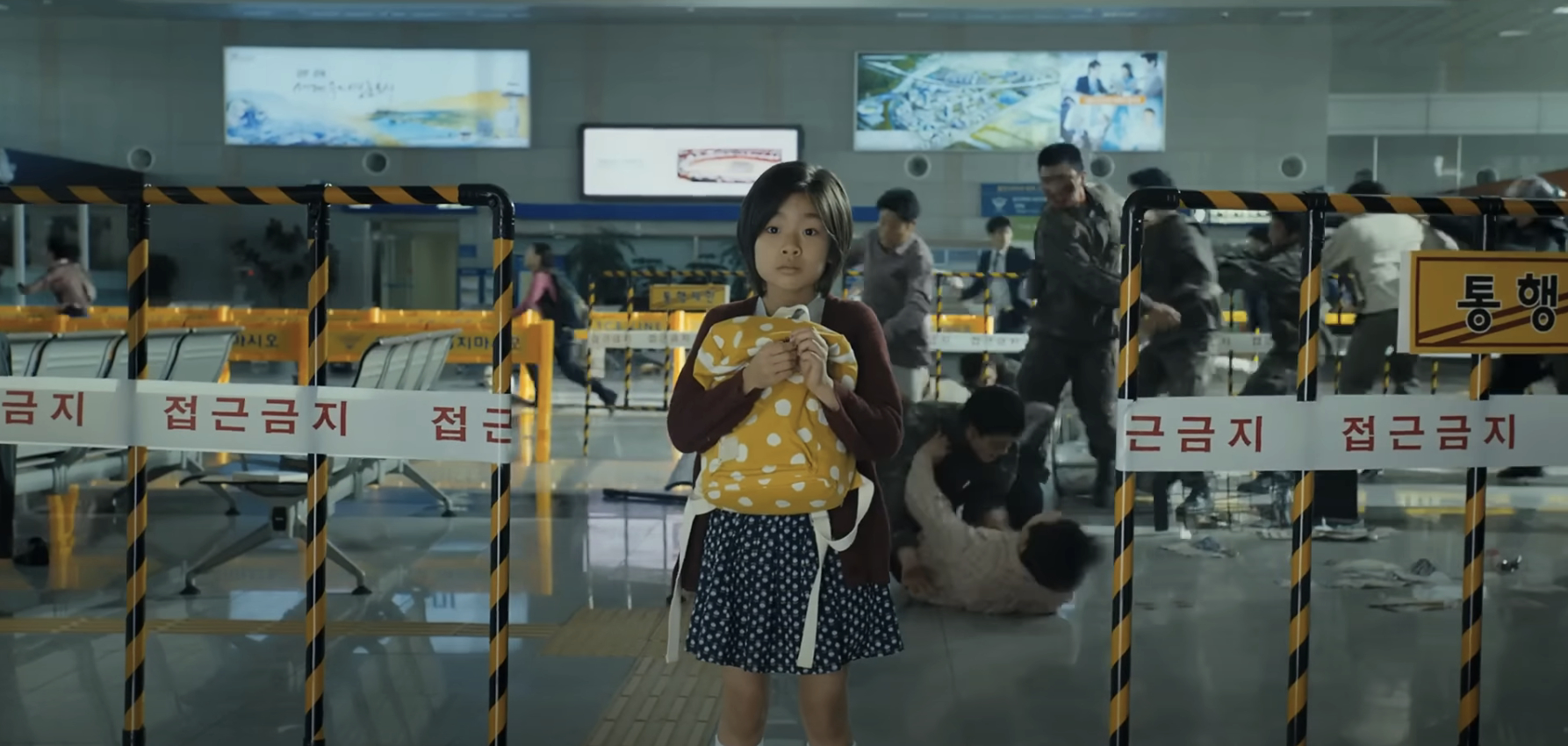 A young girl stands in an airport holding a stuffed animal. Behind her, people appear to be in chaos and distress