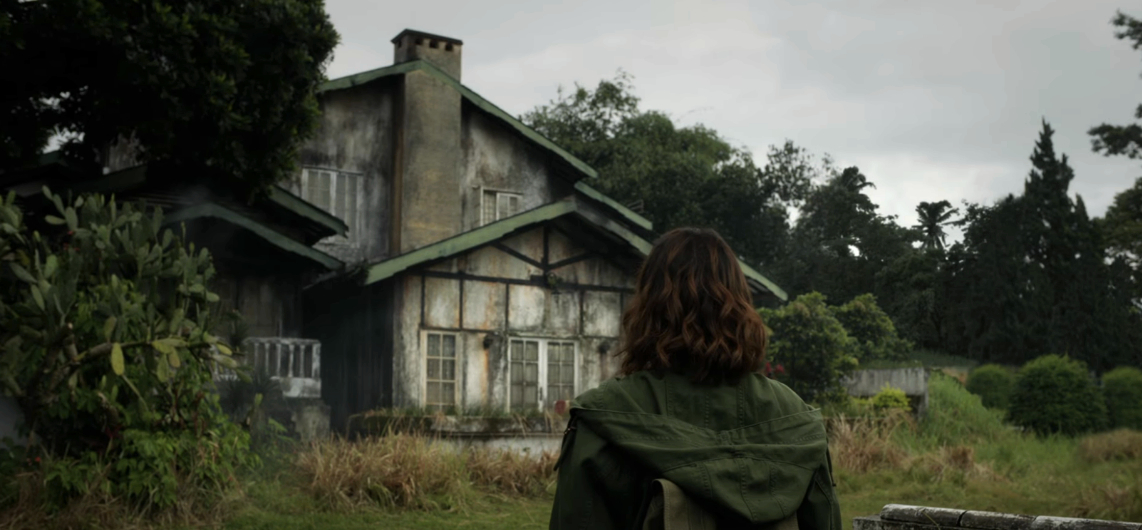 A person with long hair, wearing a jacket, stands facing an old, weathered house surrounded by greenery