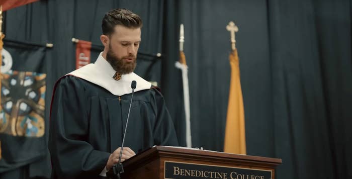 A man in graduation robes speaks at a podium labeled &quot;Benedictine College&quot; at a ceremony