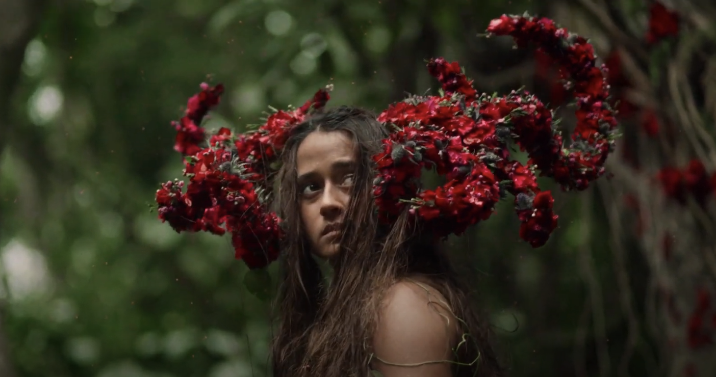 Mainei with long, loose hair looks over her shoulder while wearing a unique headpiece made of flowers in a natural, forested setting