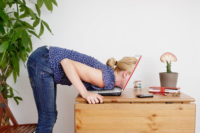 A woman leans over a laptop with her face almost touching the screen. A potted plant, cactus, and notebooks are on the wooden table next to her