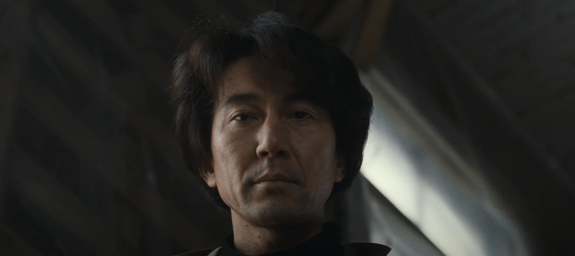 A man with medium-length hair looking solemnly downward, from a low angle, in an indoor setting