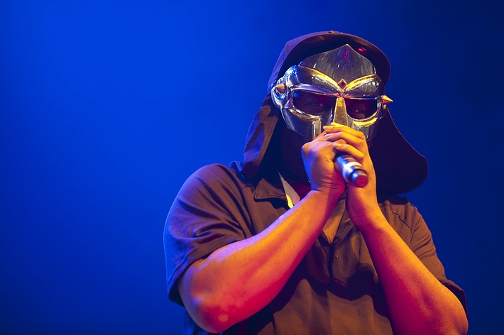 A performer, MF DOOM, is on stage wearing a metallic mask and holding a microphone. He is dressed in a dark-colored shirt with a hood