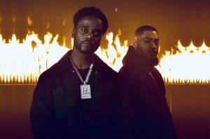 Two men stand against a wall of fire. One wears a large chain with a rectangular pendant. The other has a trimmed beard and wears a dark jacket