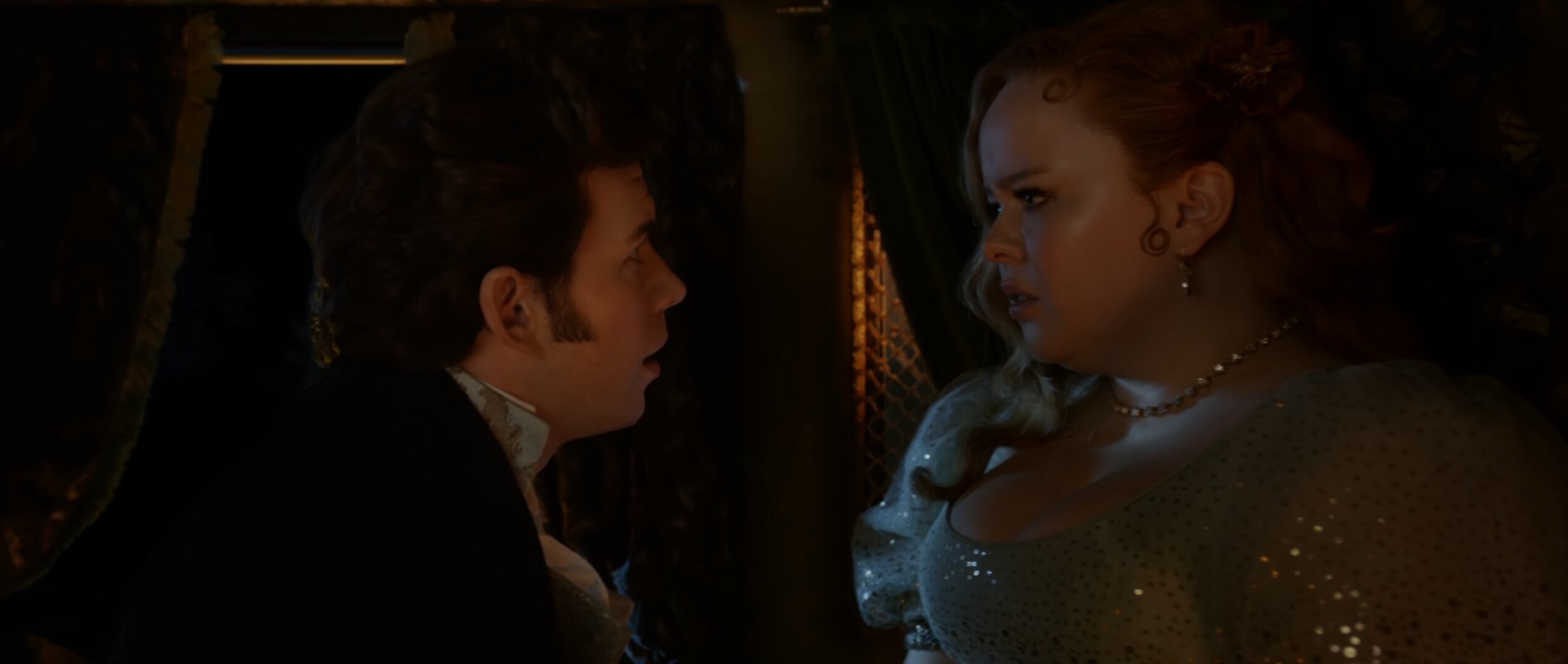 Nicola Coughlan y Luke Newton en una escena dramática de epoca, visten trajes de época en un ambiente oscuro y elegante