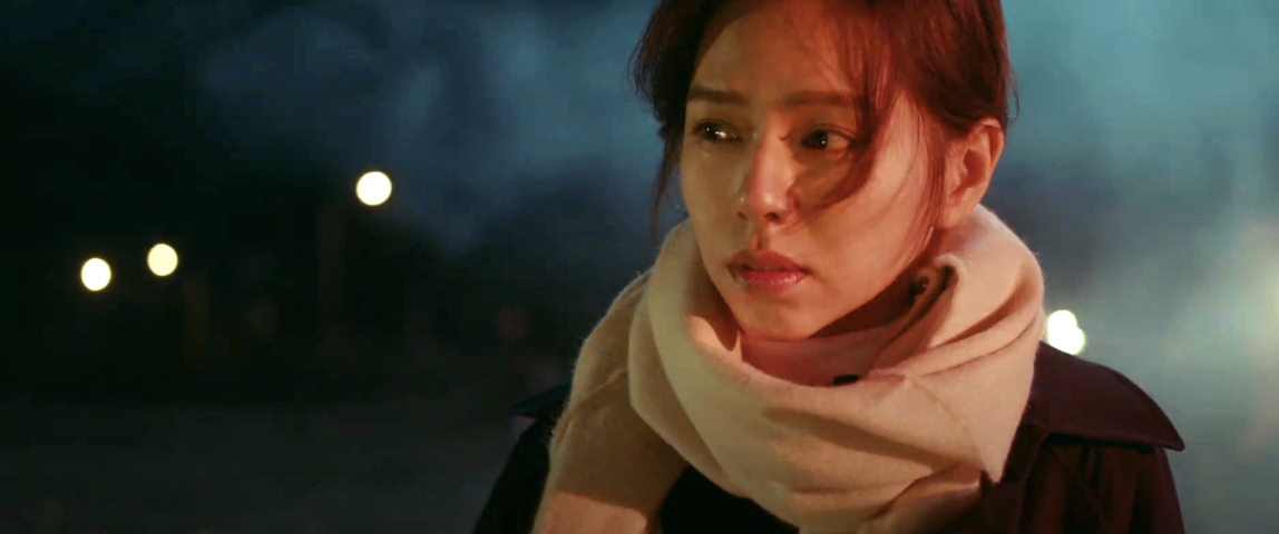 A woman with a scarf stands outdoors at night, looking contemplative, with lights and smoke in the background. No names are provided in the request
