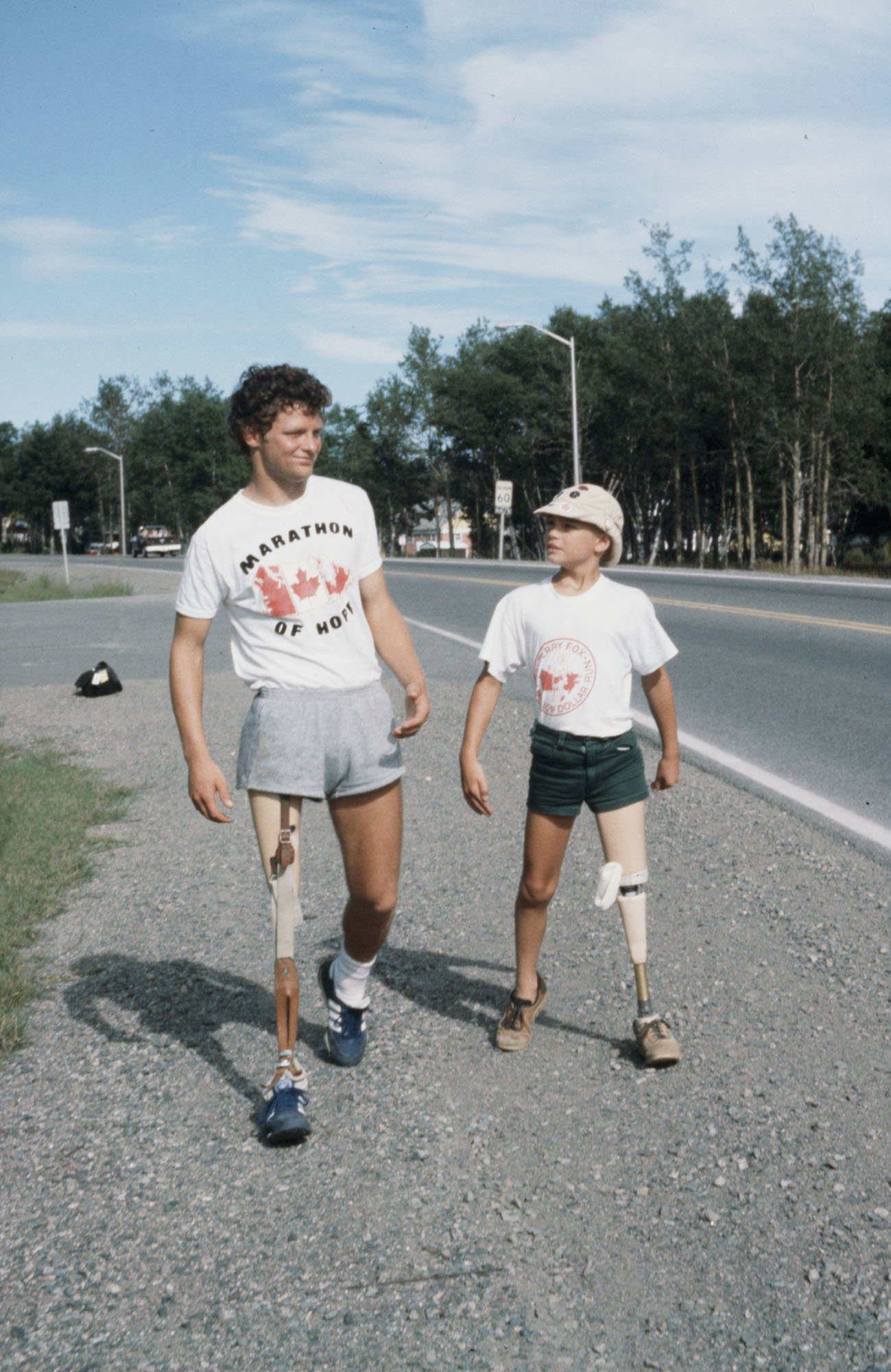Terry Fox and a young boy, both with prosthetic legs, walk along a roadside wearing casual athletic clothing