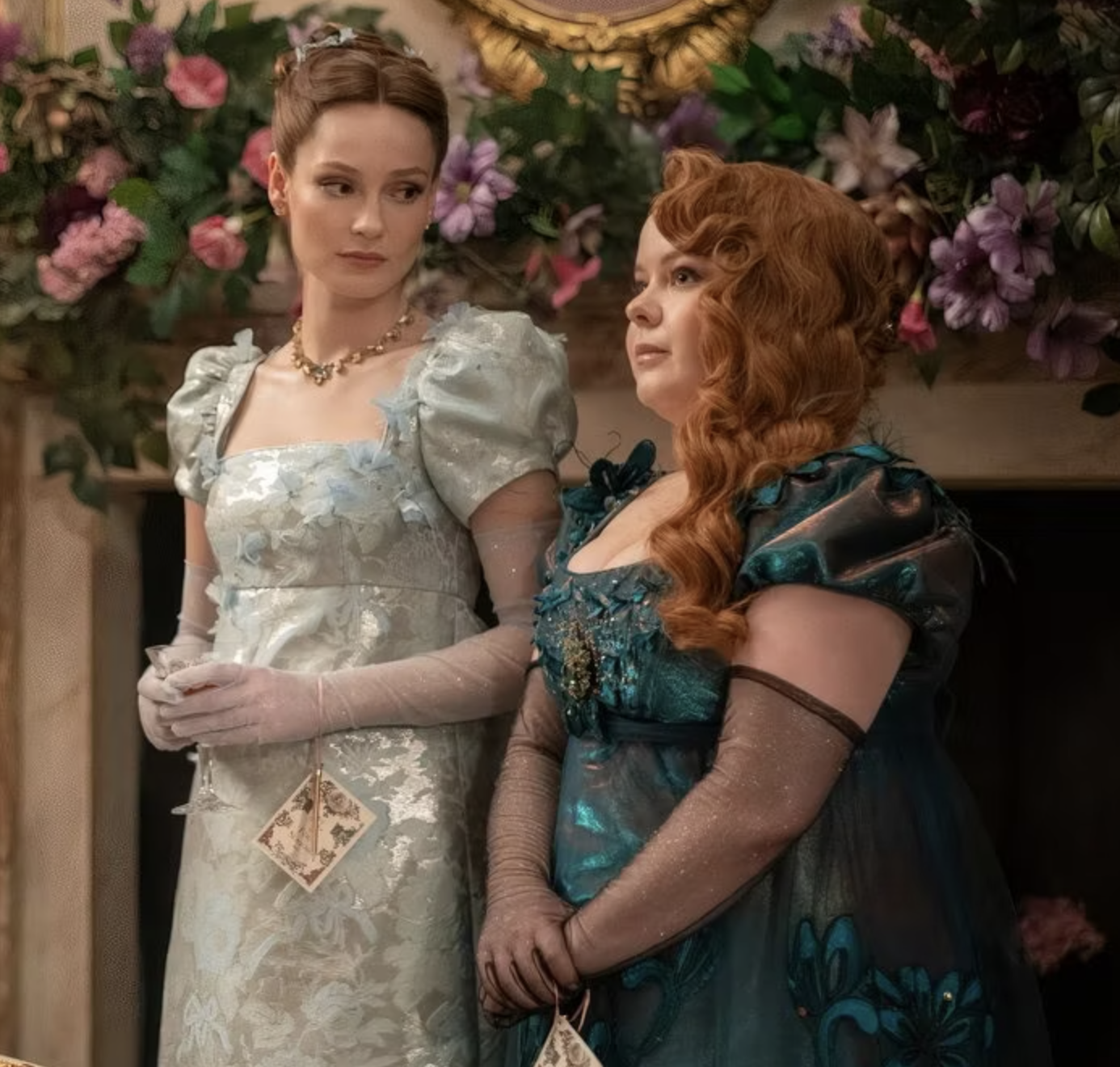 Nicola Coughlan y otra actriz en traje de época de estilo del siglo XIX en una escena de Bridgerton, rodeadas de flores