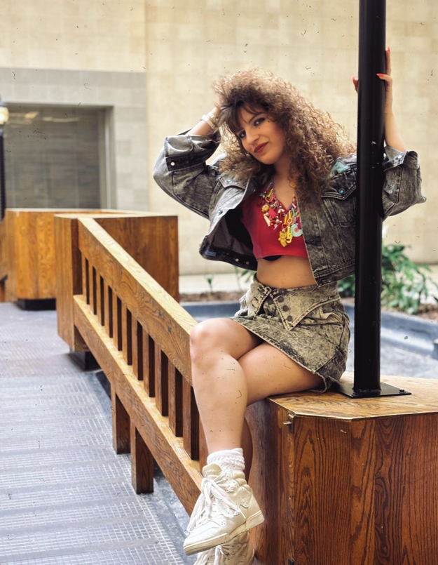 A woman with curly hair, wearing a denim jacket, crop top, and matching denim skirt, sits on a wooden railing indoors, leaning against a pole