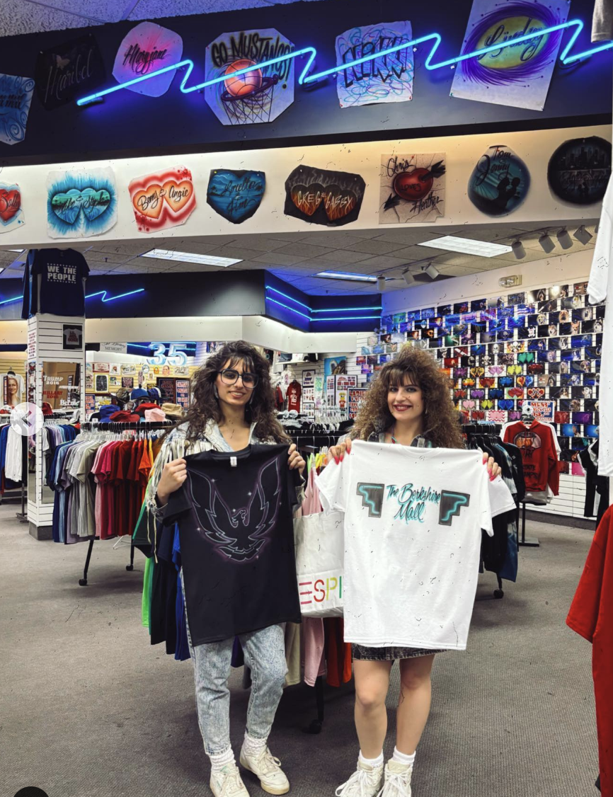 Two people in a store displaying T-shirts; walls covered with neon signs and graphic tees in the background. Names: unknown