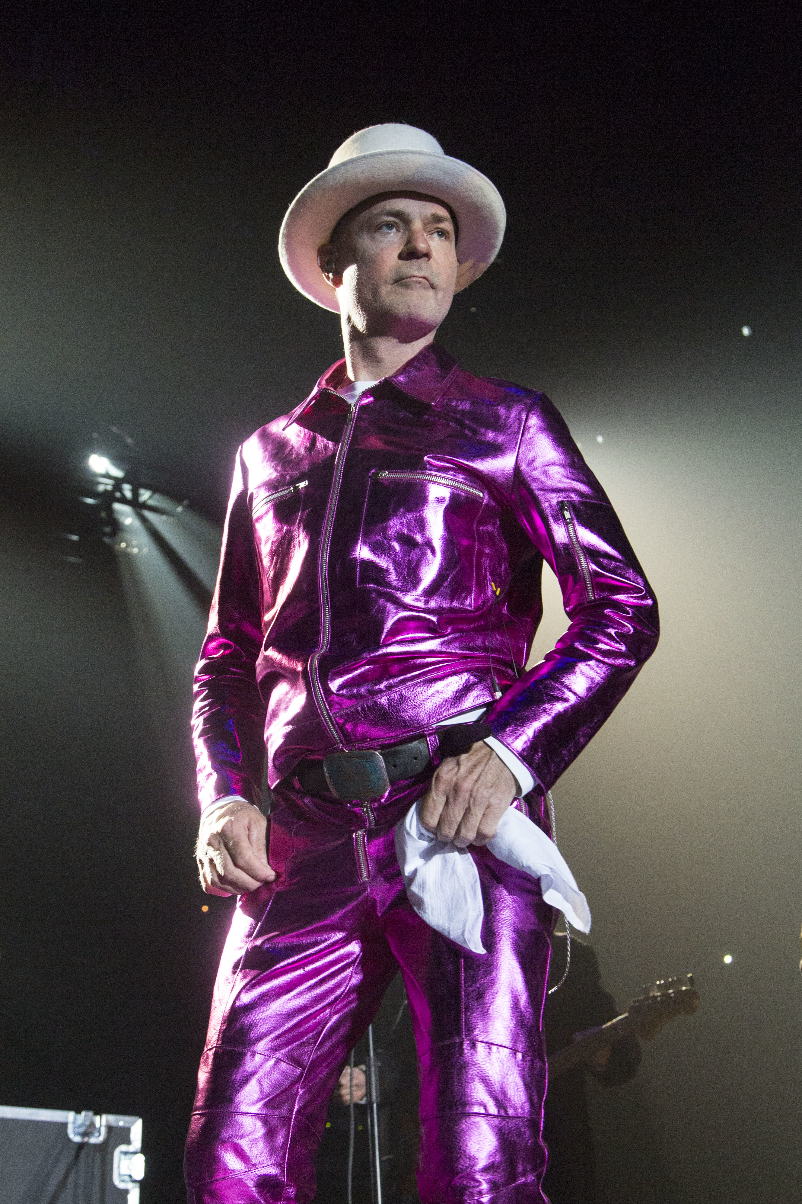 Man in a shiny purple suit and white hat holding a white cloth, standing on stage with a spotlight behind him
