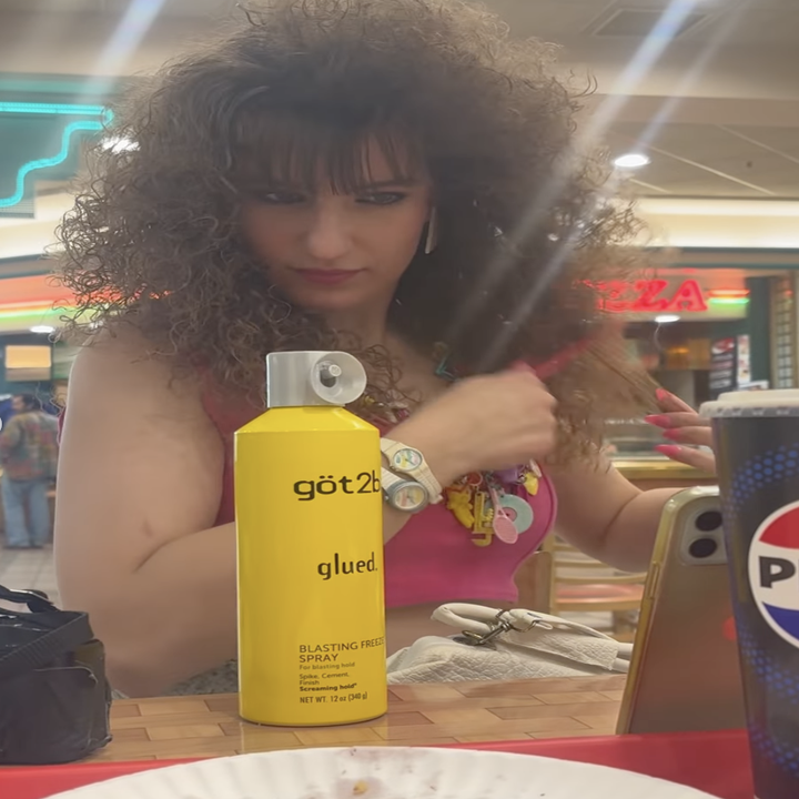 Woman at a table styling her hair with got2b glued hairspray. She's wearing a pink crop top and colorful wristbands. A Pepsi cup is visible in the foreground
