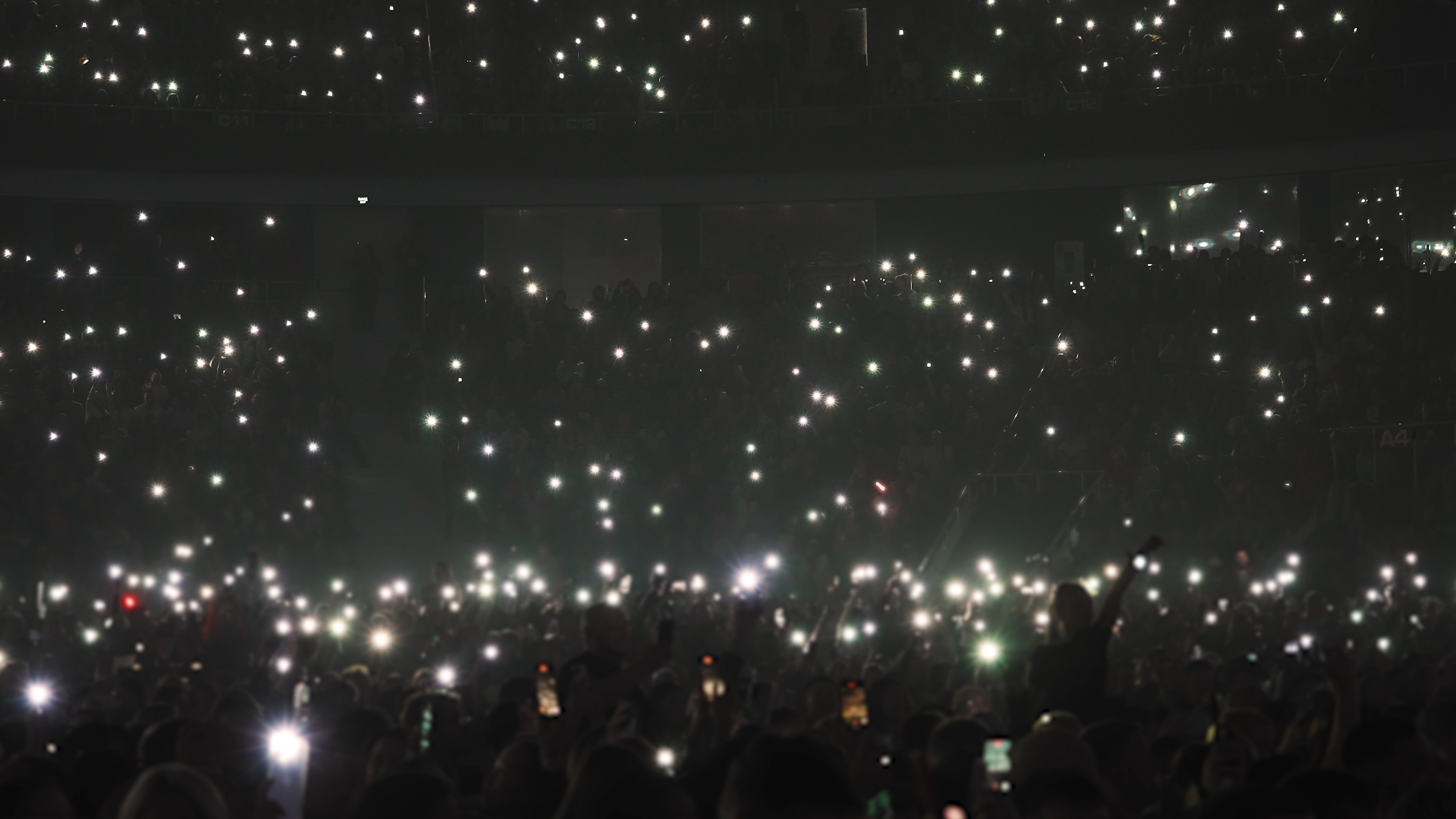 A crowd at a concert is holding up illuminated phones, creating a sea of lights in the darkened venue; the stage or performers are not visible