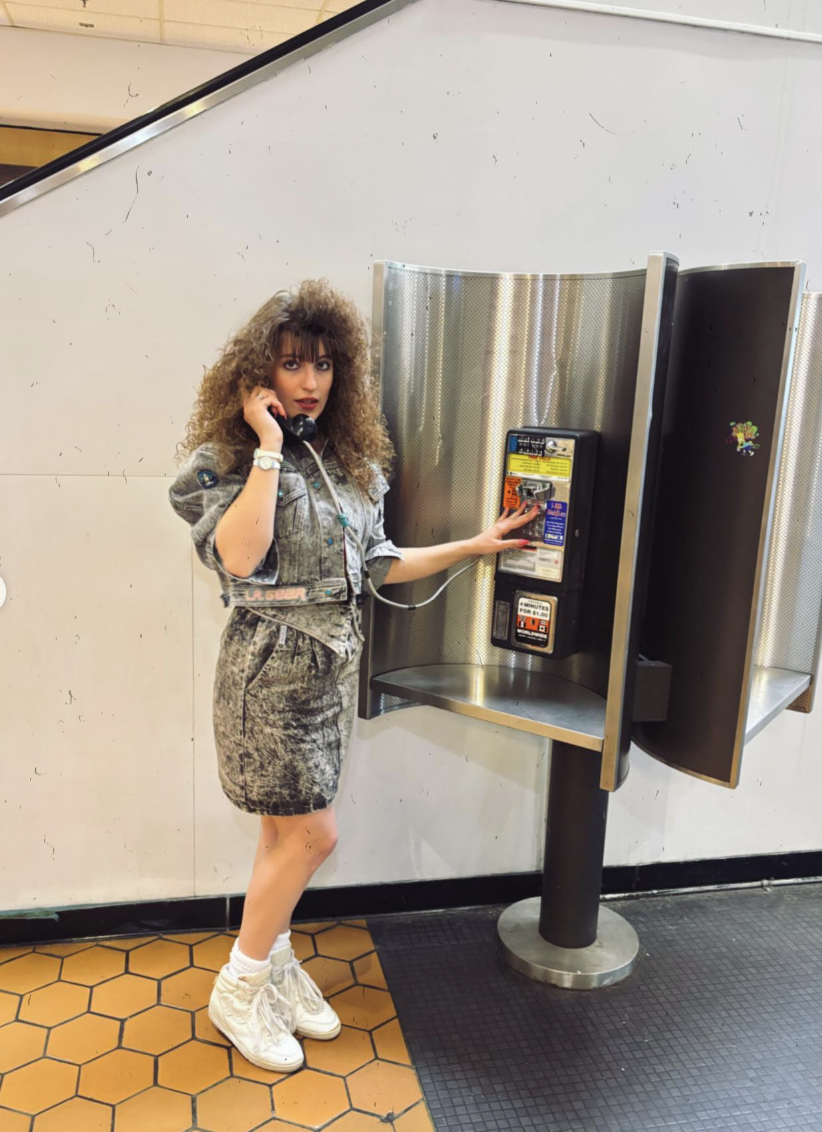 A person with curly hair is talking on a payphone, dressed in an 80s-style acid-wash denim ensemble and white high-top sneakers