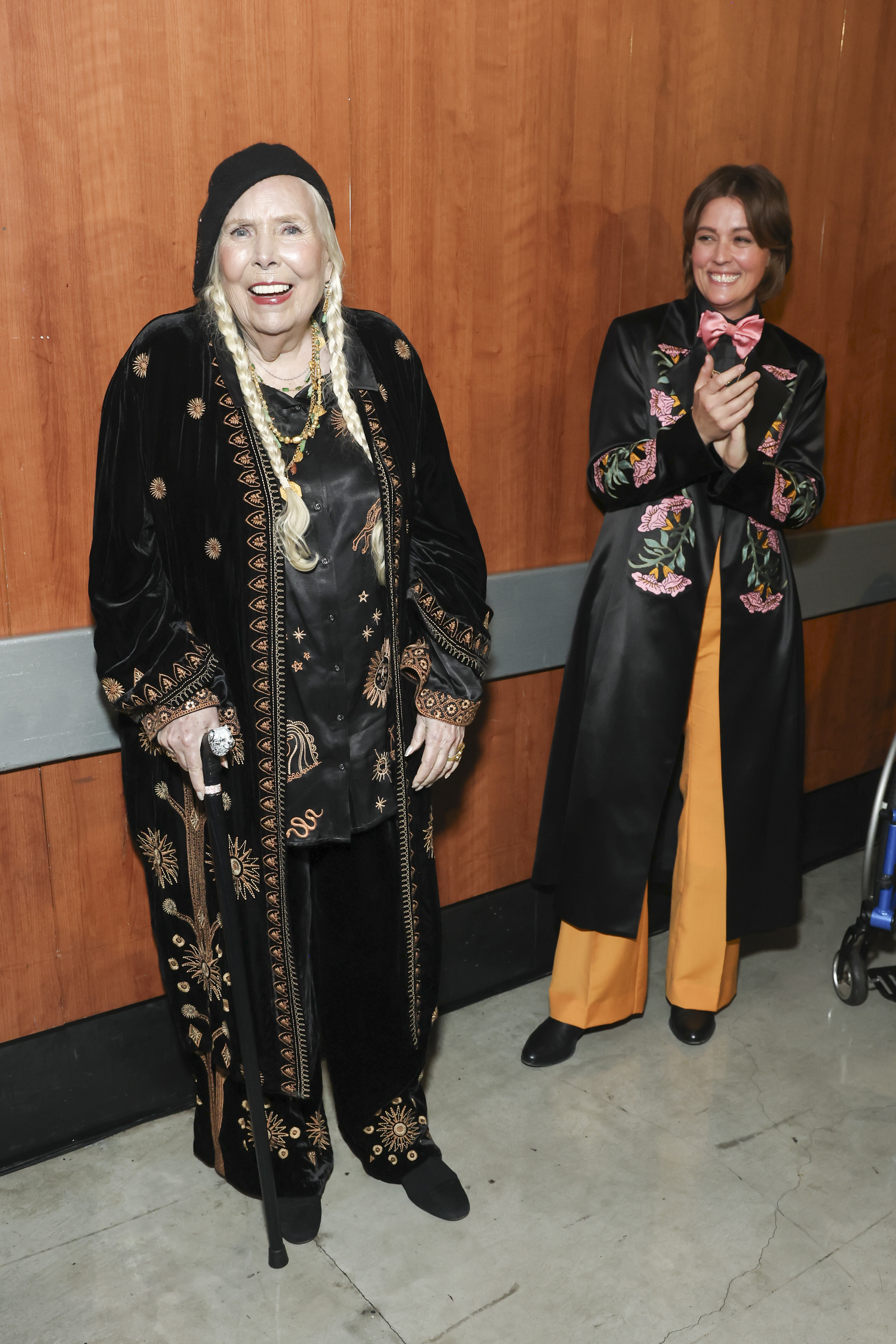 Joni Mitchell, in an ornate black outfit with gold designs and a hat, stands with a cane next to Brandi Carlile, who wears a black coat with floral embroidery and yellow pants