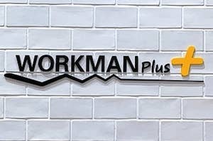 Sign reading "WORKMAN plus+" on a white brick wall above an information stand