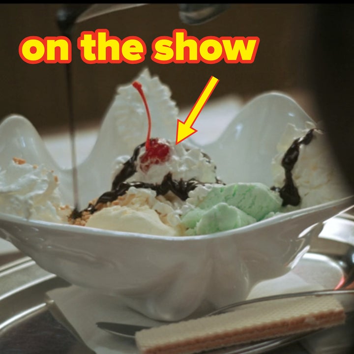 A sundae in a white dish being topped with chocolate syrup. The sundae includes whipped cream, a cherry, and a wafer on the side, placed on a silver tray