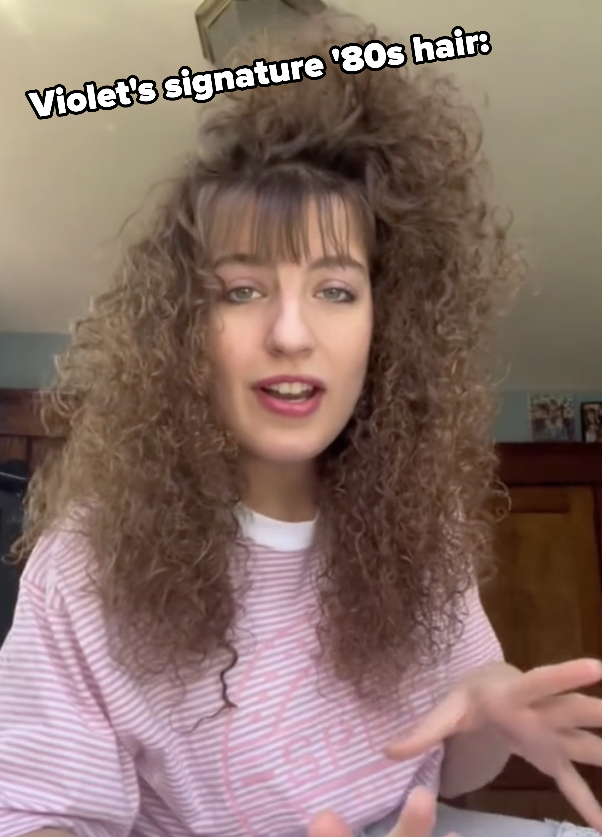 Woman with long, curly hair and bangs in a striped shirt gestures while speaking indoors