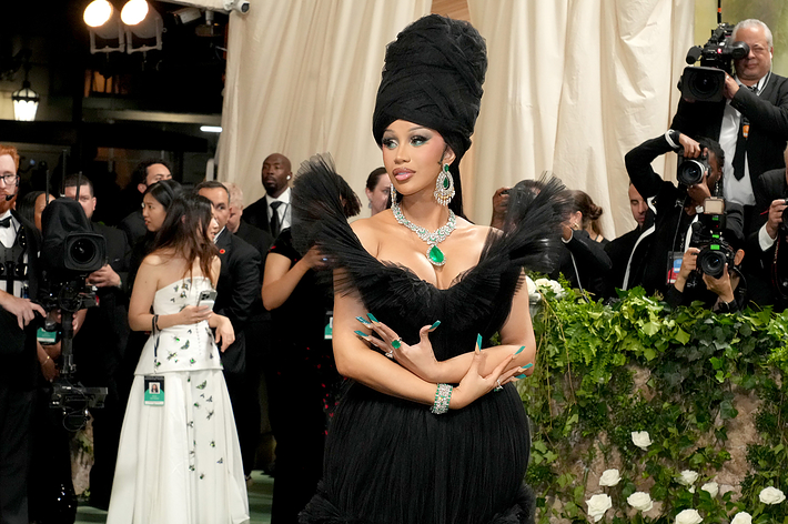 Cardi B, dressed in an elegant black gown with dramatic ruffles and a tall headpiece, stands on the red carpet at a glamorous event surrounded by photographers