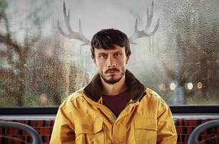 Richard Gadd in a yellow jacket sits in front of a window with rain and deer antler reflections