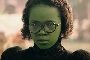 Person with a green face, wearing round glasses and a black, long-sleeve, high-neck dress in a serious expression. Name unknown