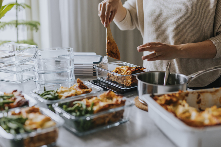 Person in a kitchen preparing multiple containers of lasagna and green beans with a wooden spatula
