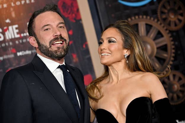 Ben Affleck in a suit and tie and Jennifer Lopez in a strapless gown smile together at an event, with a background featuring gears and a sign