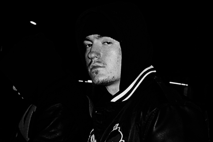A man wearing a hooded jacket and a beanie at night. He has a serious expression and is looking towards the camera. His identity is not known