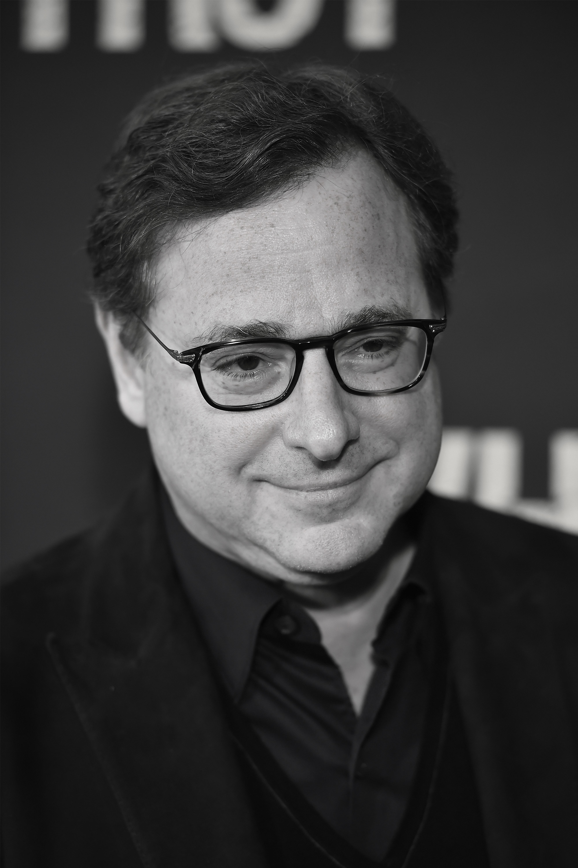Bob Saget at a public event, wearing glasses and a suit with an open-collared shirt