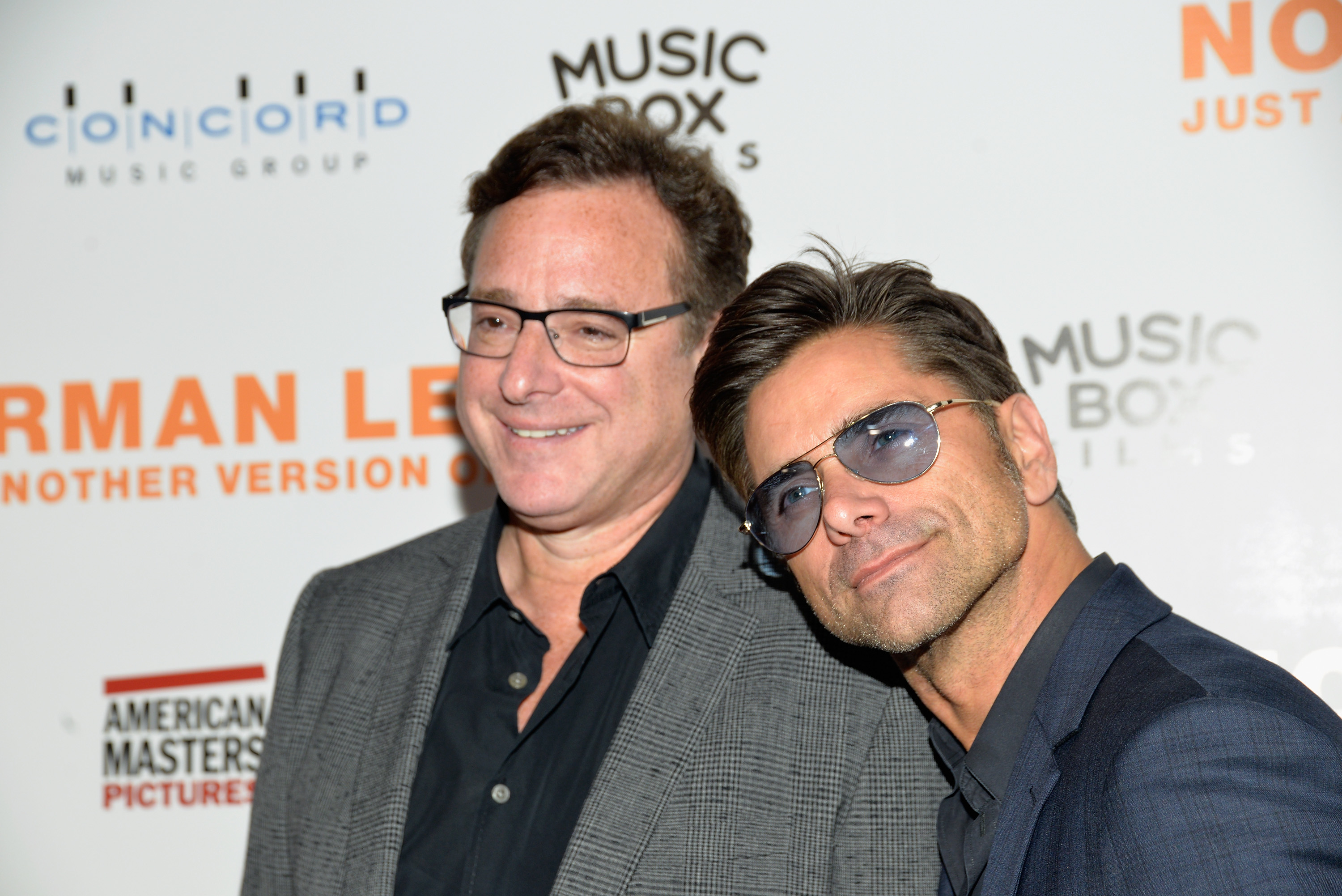Bob Saget and John Stamos at a Music Box Films event, smiling for the camera. Saget wears a suit jacket over a shirt, while Stamos wears a blazer with sunglasses