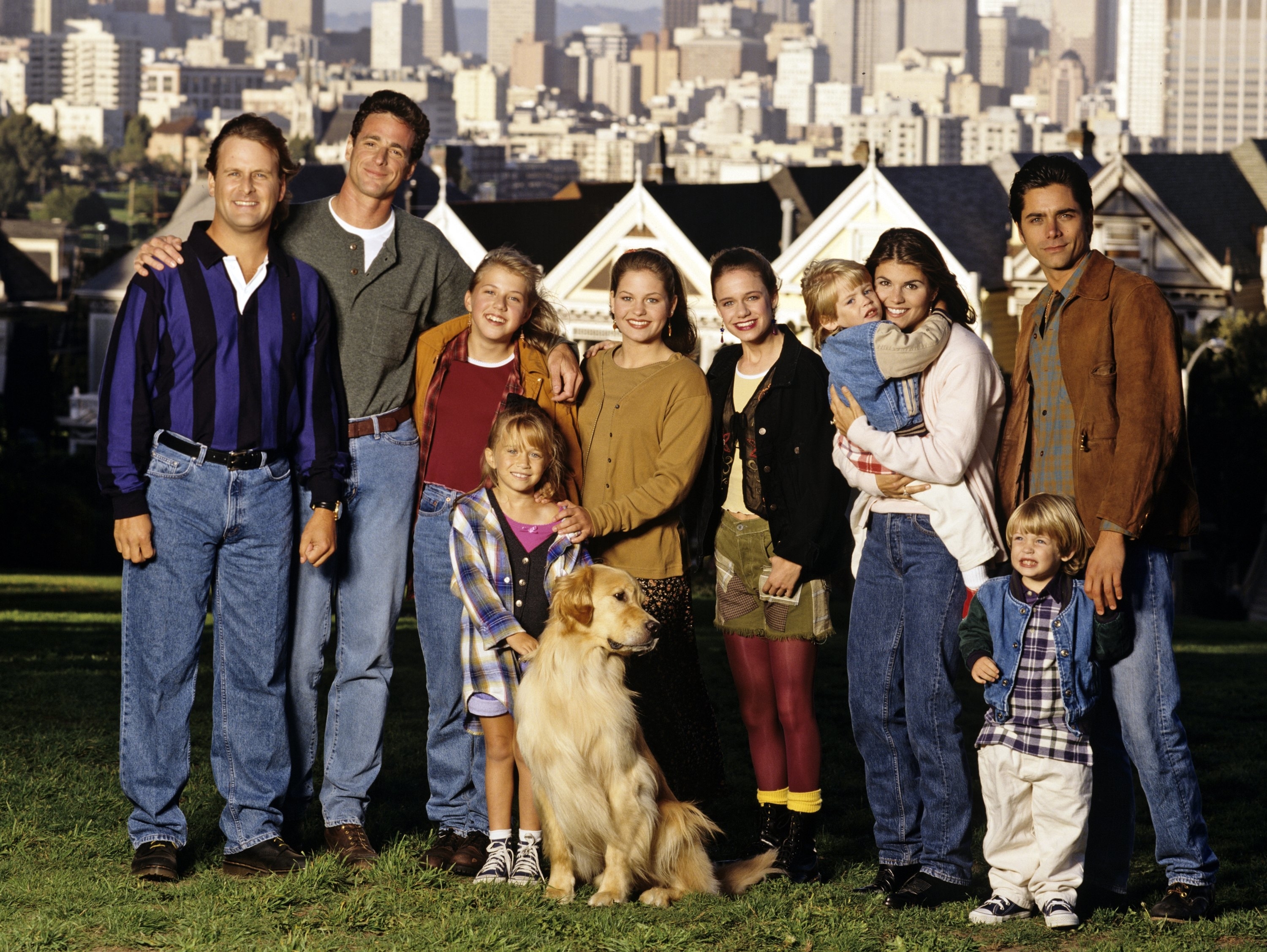 The image shows the cast of &quot;Full House&quot; posing outdoors with the San Francisco skyline in the background. Members include Bob Saget, Dave Coulier, John Stamos, and others