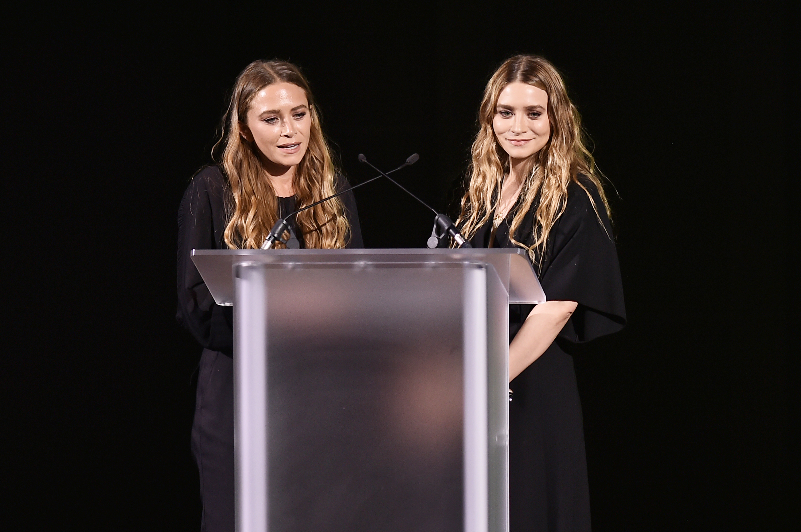 Mary-Kate and Ashley Olsen stand at a podium, dressed in black outfits, possibly delivering a speech or presentation