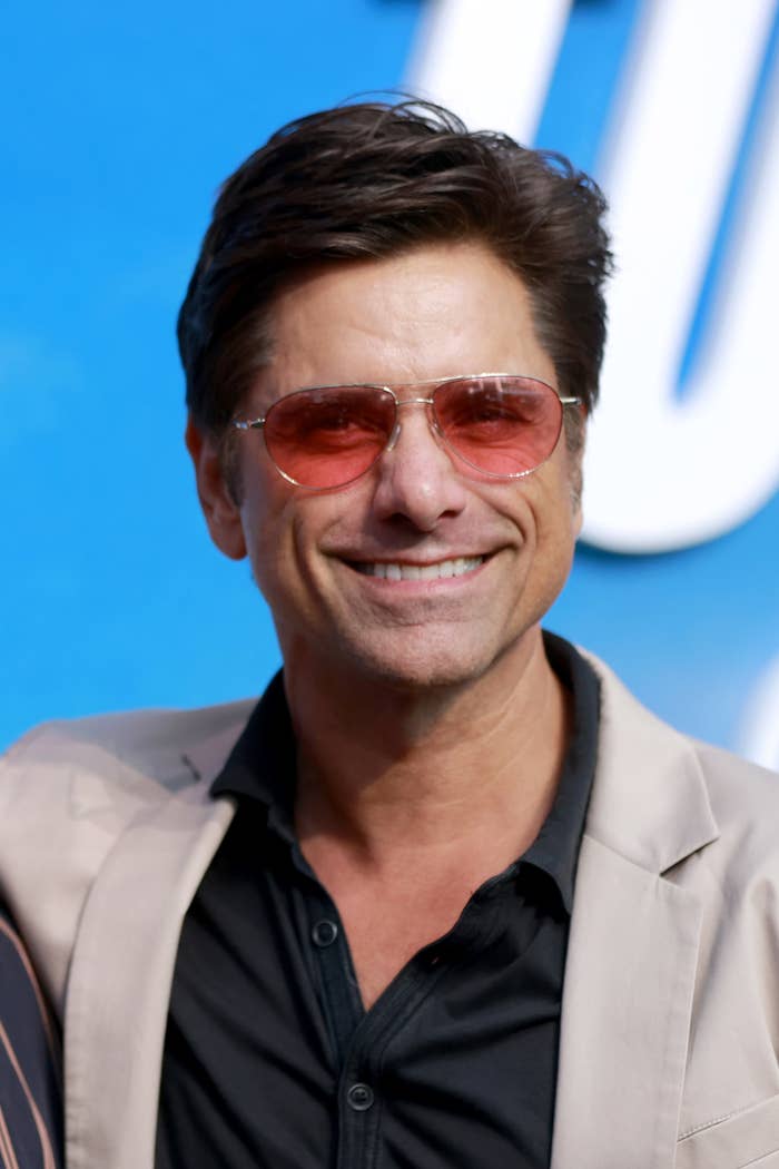 John Stamos smiles while wearing a beige jacket, black shirt, and pink-tinted sunglasses at a public event