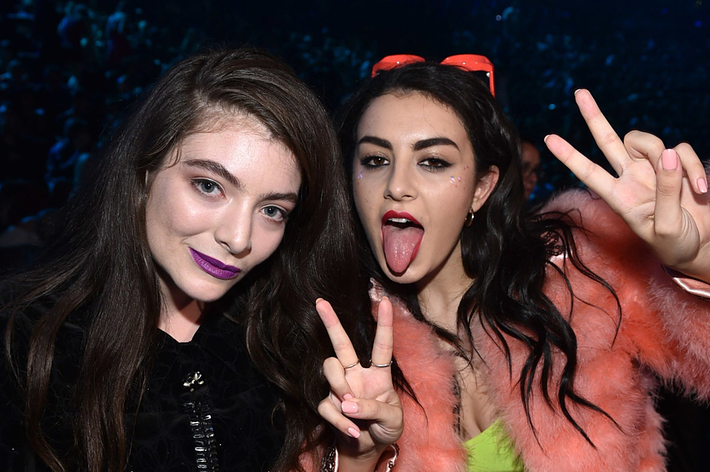 Lorde and Charli XCX make peace signs at a music event. Charli wears a fur jacket and sunglasses, while Lorde is in a dark top